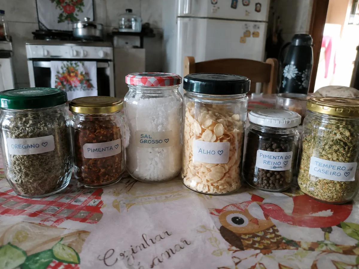 Fileira de potes de vidro com orégano, pimenta, sal grosso, alho e tempero caseiro